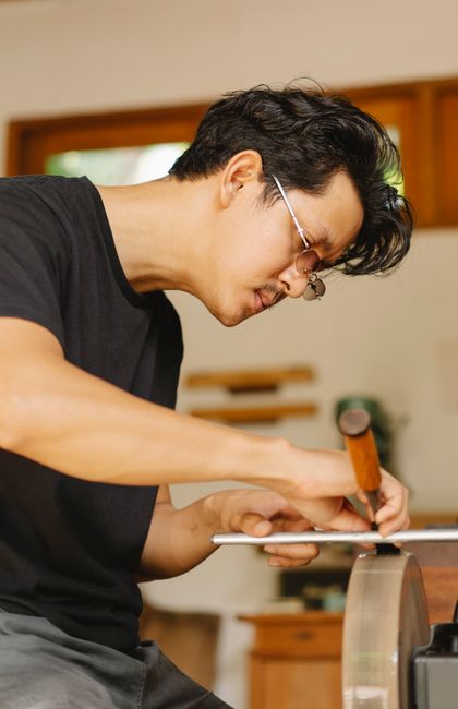 A man with dark hair and glasses, wearing a black T-shirt, is inside a home preparing for a move. He is standing near a wooden countertop, using a hammer and a metal tool to assemble or disassemble furniture or packing materials. The background shows kitchen cabinets and a window that allows natural light into the room. The workspace is organized, and the man appears focused on his task, which is part of a home relocation or packing process. This scene exemplifies key stages in furniture assembly or disassembly during house removals, highlighting the attention to detail required in professional moving services offered by Man with Van Whetstone, supporting the logistical aspects of packing and furniture transport.