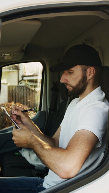 A man with a beard, wearing a black cap and a white polo shirt, sits inside a van with a light-colored interior, focused on using a smartphone. The background visible through the windshield shows an outdoor area with a wooden fence and some greenery. The man appears to be part of a home relocation process, possibly coordinating or checking details during loading or planning of furniture transport with Man with Van Whetstone, a professional removals service. The van's interior indicates a typical vehicle used for house removals, with space for packing materials and furniture. The lighting is natural, illuminating the man's face and the inside of the vehicle, emphasizing the calm and professional atmosphere of the moving process.