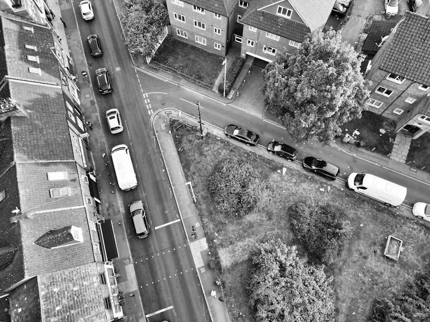An aerial black-and-white photograph captures a residential street during a home relocation process on a day with overcast lighting. Several vehicles are parked along both sides of the narrow street, including a white van positioned near the curb at the front right of the image, which is likely used for furniture transport during the move. In front of a row of terraced houses with pitched roofs and brick facades, a section of the pavement shows a clutter of cardboard boxes, plastic-wrapped furniture, and wrapped packages, indicating packing and moving activities. A few large pieces of furniture, such as a wooden table or wardrobe, are visible near the entrance of one property, with some items partially placed inside the house doorway ready for loading. Moving equipment like trolleys or straps is not visible, but the scene involves the loading process onto the van parked on the street. The surrounding area features trees and landscaped patches of grass, with a small public garden or park area adjacent to the houses, and a street with traffic markings and curbside spaces for parking, supported by Man with Van Whetstone's relocation services, reflecting a typical scheduled move in the N20 postcode area, potentially involving a local house removal or property transfer.