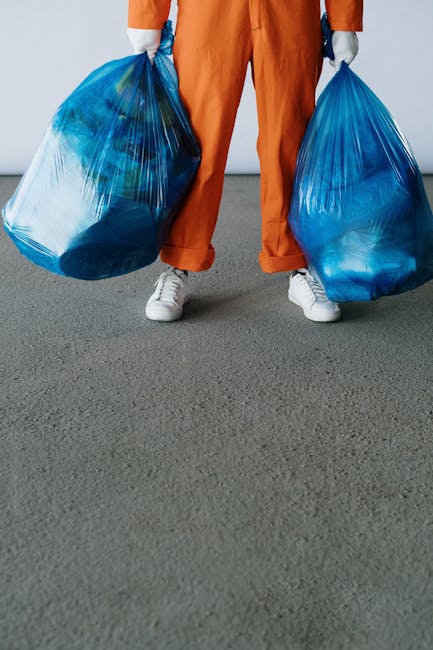 A person standing indoors on a grey carpeted floor, holding two large blue plastic rubbish bags filled with household waste. The individual is wearing orange work trousers, white sneakers, and grey gloves, with only their lower body visible in the image. In the background, a doorway leads to a room with a wooden door frame, and the lighting suggests natural or diffused indoor illumination. The scene depicts the process of waste collection or disposal, which may be related to a home relocation or clearance service provided by Man with Van Whetstone, highlighting the distinction between bulky waste removal and professional house removals. The bags are positioned slightly apart, with one in each hand, indicating an effort to load or carry waste items for transport, possibly onto a vehicle for disposal or recycling. The setting emphasizes a clean, organized environment suitable for household waste management as part of moving or clearance activities.