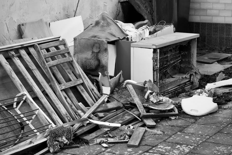 The image depicts an outdoor area with an assortment of discarded household items and debris, including wooden furniture such as a dismantled wooden chair and a small wooden pallet, along with broken pieces of wood and debris scattered on the paved ground. There is a white plastic object, possibly a container or small appliance, lying on the pavement, and various cardboard and paper materials are also present. Behind these objects, there is a partially open doorway revealing an indoor space, with a damaged appliance or piece of furniture partially visible inside, covered with a cloth or protective wrapping. To the right, a brick wall is visible, and the overall scene appears to be the aftermath of clearing or rubbish disposal related to house removals or renovation waste, which a professional removals service like Man with Van Whetstone might handle during a home relocation or clearance process.