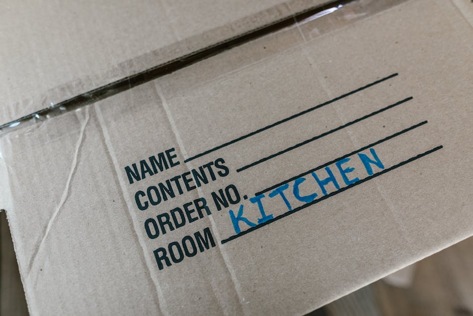 Close-up of a cardboard moving box with black printed labels for packing and home relocation, showing handwritten blue text indicating 'ROOM KITCHEN' for the designated contents. The box is sealed with clear packing tape and positioned on a flat surface inside a property, likely preparing for furniture transport or loading into a van. The background reveals other boxes and packing materials, suggesting an ongoing packing or moving process typical of house removals handled by Man with Van Whetstone. The scene emphasizes careful packing organization for a home move, with natural indoor lighting highlighting the label and materials used for protective packaging during furniture transport and loadings.