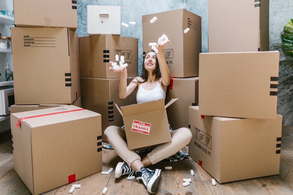 A young woman with long dark hair sitting cross-legged on a wooden floor inside a home surrounded by multiple cardboard boxes of various sizes, some sealed with red tape and others open revealing packing materials such as white foam packing peanuts. She is wearing a white sleeveless top, beige pants, and black and white sneakers, smiling as she tosses foam peanuts into the air. The boxes are stacked against a textured blue wall, with a white microwave visible on a countertop to the left and a green potted plant on the right side of the image. This scene depicts the packing process involved in a home relocation, with the woman preparing to move. The setting is well-lit with natural light, capturing the typical logistics of furniture transport and packing associated with house removals, as could be handled by companies like Man with Van Whetstone during residential moves.