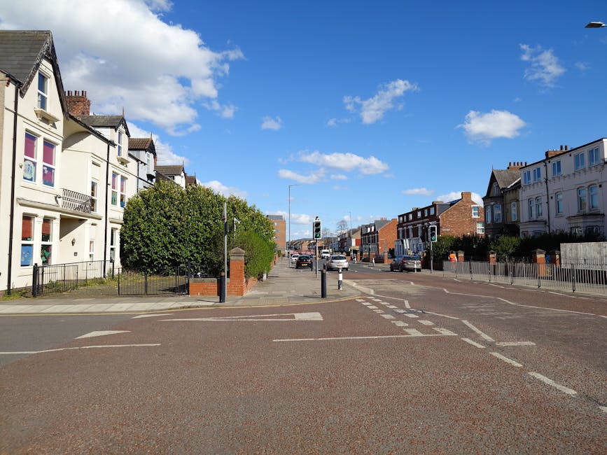 A wide view of Oakleigh Road North in Whetstone on a bright day with blue sky and scattered clouds. On the left, a row of light-colored, multi-story residential buildings with bay windows, small balconies, and decorative architectural details are visible. In front of these buildings, there is a small pavement area separated by a low metal fence, with a large, neatly trimmed green tree near the corner of the street. On the right, additional residential properties with similar styles are seen, bordered by low fences and small front gardens. The road itself features typical urban markings, including pedestrian crossings and lane divider lines, with a few cars stopped at traffic lights in the distance. The traffic lights are mounted on black poles with traffic control signals visible. Overall, the scene depicts a calm suburban street suitable for home relocations or furniture transport logistics associated with house removals, as represented by the context of relocation services on manwithvanwhetstone.co.uk.