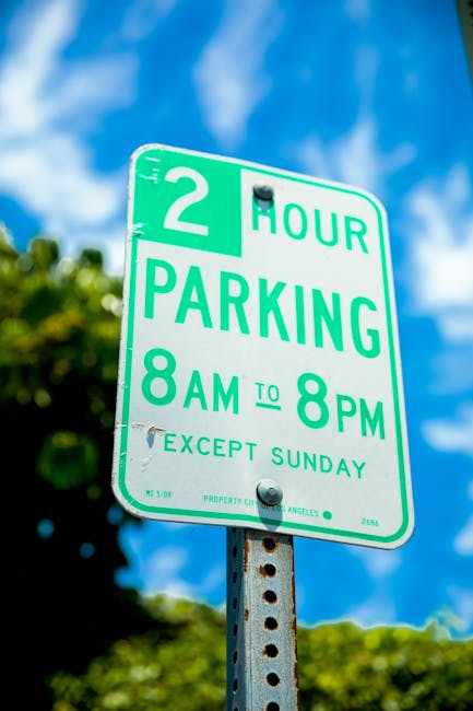 A green and white parking restriction sign mounted on a metal pole outdoors, indicating a two-hour parking limit from 8 am to 8 pm, except on Sundays. The sign is located in a residential or urban are