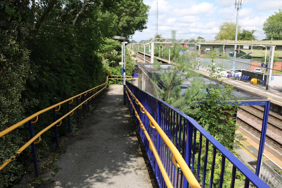 A narrow outdoor footpath at Totteridge & Whetstone station featuring a black asphalt surface with shadows cast by nearby trees, bordered on the left side by dense green foliage and on the right by a blue metal railing with yellow handrails. The pathway leads to a set of stairs or an accessible lift station entrance, which is used for home relocation and furniture transport access. In the background, railway tracks run parallel to the station platform, with overhead power lines supported by metal pylons, and parked cars visible beyond a security fence. The station platform has a yellow safety line along its edge, with some benches and small structures visible on the far side. The scene is illuminated by natural daylight under a partly cloudy sky, indicating daytime conditions suitable for moving and packing activities. Man with Van Whetstone occasionally utilizes such station access points for efficient relocation logistics.