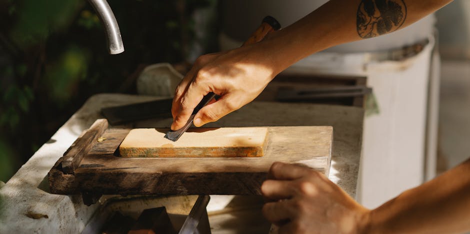 Close-up image of a person's hands during a packing and moving process, with one hand holding a small chisel and the other stabilising a weathered wooden board, which is positioned on a larger wooden surface indoors. The person is wearing a short-sleeved shirt, revealing tattoos on the arms, and the workspace is illuminated by natural light. The background shows a blurred kitchen or utility area, with items such as a white appliance and some greenery visible. This scene captures an element of furniture preparation or disassembly in the context of house removals, relevant to professional relocation services provided by Man with Van Whetstone, illustrating the careful handling and packing of household items for a home relocation or furniture transport.