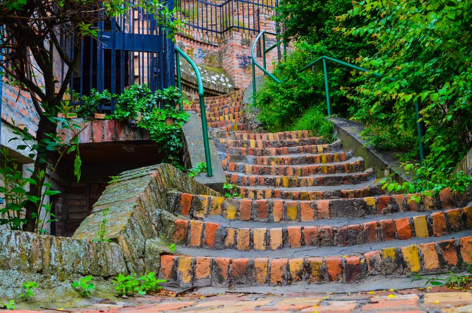 A set of outdoor brick stairs with uneven, weathered steps leading upward in a residential area. The stairs are bordered by green metal handrails on both sides and are surrounded by lush green foliage, including bushes and small trees. The brickwork shows signs of age and wear, with some moss and dirt visible on the steps. To the left is a section of brick wall partially covered with plants, and the stairs are part of a home relocation or moving process, possibly being prepared for furniture or boxes to be carried up or down, with a gravel or paved pathway at the base. The background includes a black metal fence and additional brick structures, with natural lighting illuminating the scene, highlighting the textures of the bricks and greenery. This image, associated with Man with Van Whetstone, reflects the logistical considerations during house removals involving outdoor staircase access.