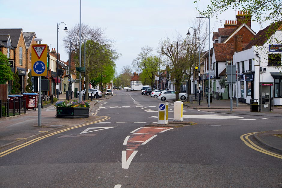 A wide street in a residential and commercial area with a central roundabout and traffic calming measures, including bollards with reflective markings. On the left side, there is a row of buildings wi