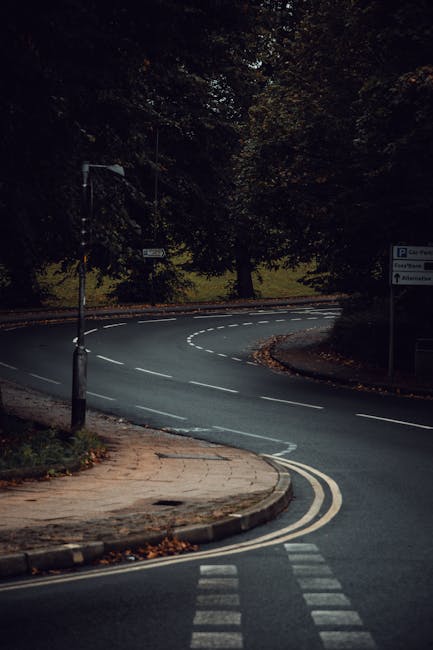 A wide street in a residential and commercial area with a central roundabout and traffic calming measures, including bollards with reflective markings. On the left side, there is a row of buildings wi