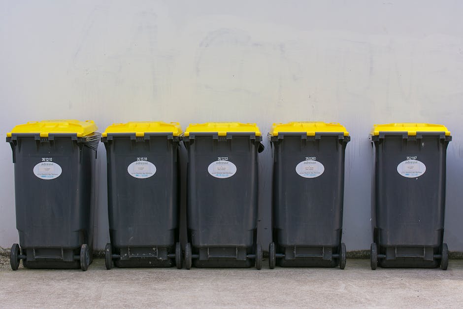 Five large wheeled rubbish bins made of dark grey plastic with yellow lids, lined up in a row outdoors against a plain light grey wall. The bins are positioned on a concrete surface, with some visible wear and dirt on their lower sections. Each bin has a white oval label with black text and barcode, indicating identification numbers. The scene appears to be part of a waste disposal area adjacent to a property, with natural daylight illuminating the scene. This arrangement suggests the bins are ready for collection as part of a home relocation or clearing process, which may be managed by a removals company like Man with Van Whetstone, specializing in furniture transport and packing services.
