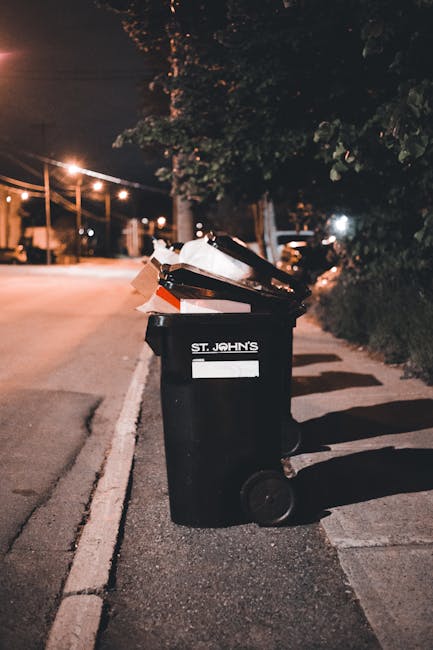 A black wheelie bin placed on a paved sidewalk beside a street at night, labeled with the words 'St. John's'. The lid is open and filled with various rubbish, including cardboard boxes and plastic packaging, some of which are spilling slightly over the top. In the background, there is a row of streetlights illuminating the road and a line of trees and bushes alongside the pavement. The scene suggests waste disposal as part of a house relocation or packing process, with the setting indicating an outdoor area ready for rubbish collection, consistent with guidelines on legal rubbish disposal during home moving services provided by Man with Van Whetstone and their removals team.